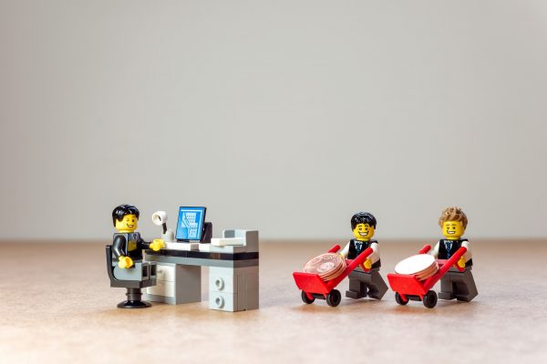 Kuvituskuva. Legohahmo istuu työpöydän takana silmää iskien, kaksi hahmoa kuljettaa nokkakärryillä kolikkoja .